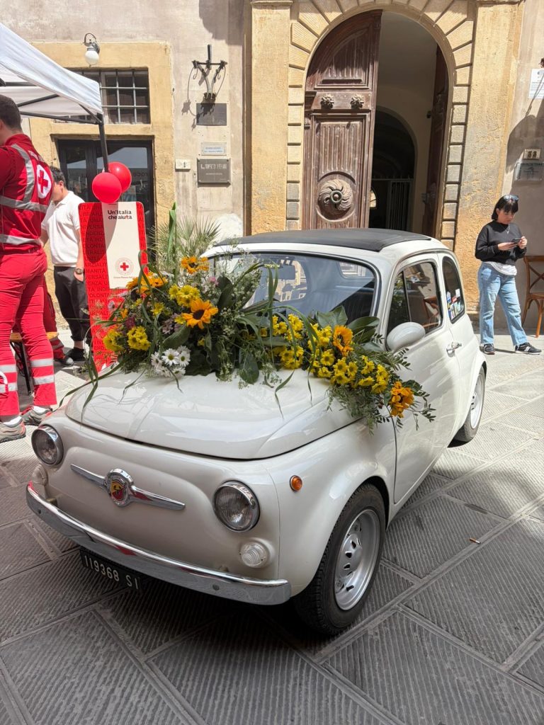 A small, white vintage FIAT car with its windshield covered in a large yellow and orange flower arrangement  